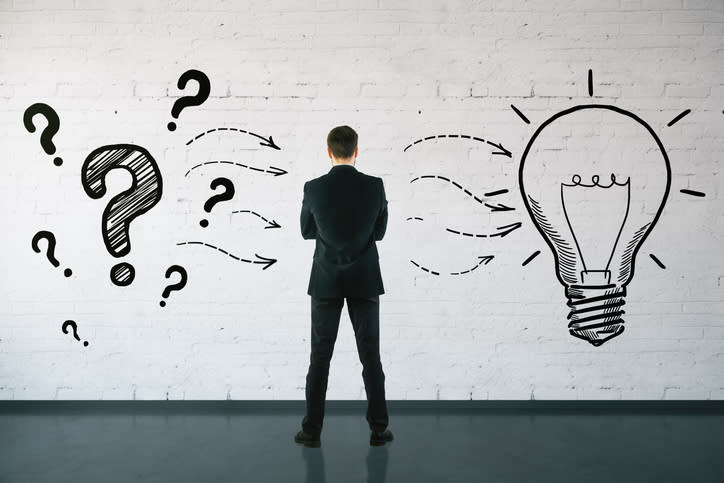 A businessman with his back to the camera staring at a drawing of question marks and a light bulb on the wall.