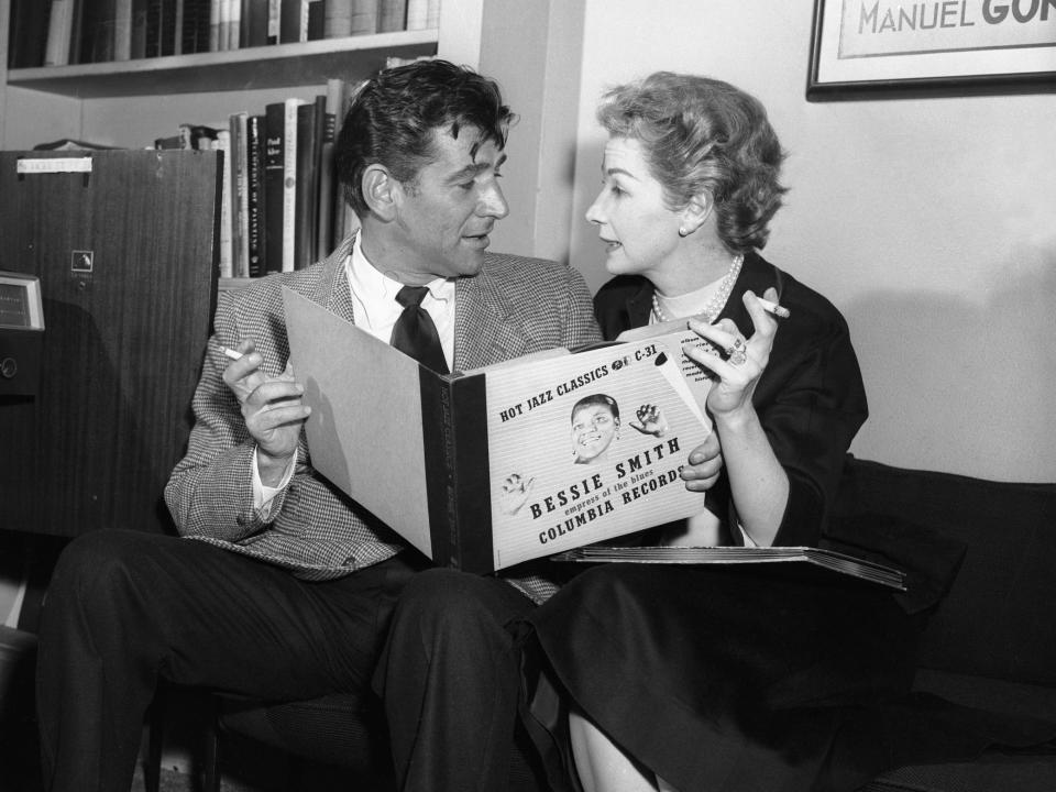 Leonard Bernstein and Felicia Montealegre sit on a couch, both holding cigarettes and a record album, in this 1956 black and white photo.