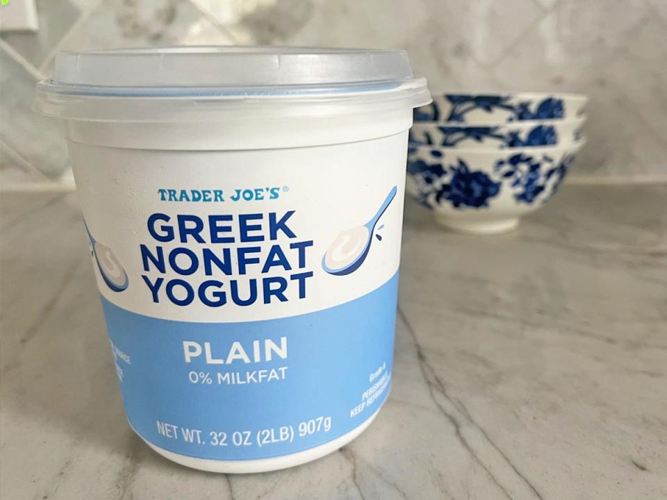 A white and light-blue tub of Trader Joe's Greek nonfat yogurt, with illustrations of spoons on the packaging. The container sits on a gray marble countertop with blue-and-white bowls in the background