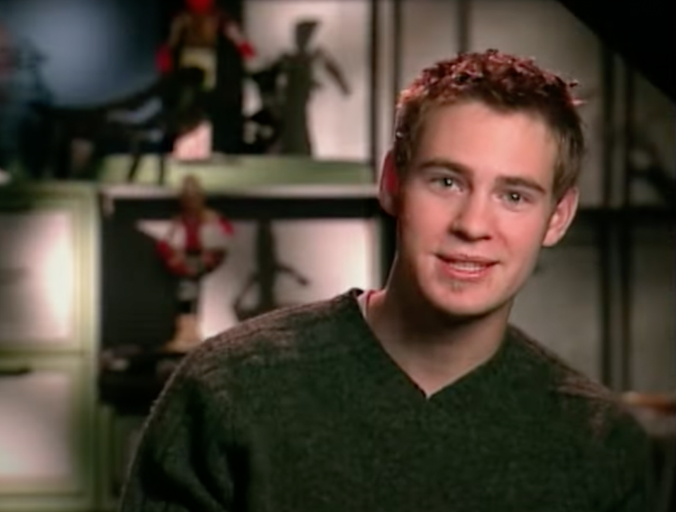 A young man with short brown hair wearing a dark sweater stands in front of shelves displaying various action figures and collectibles