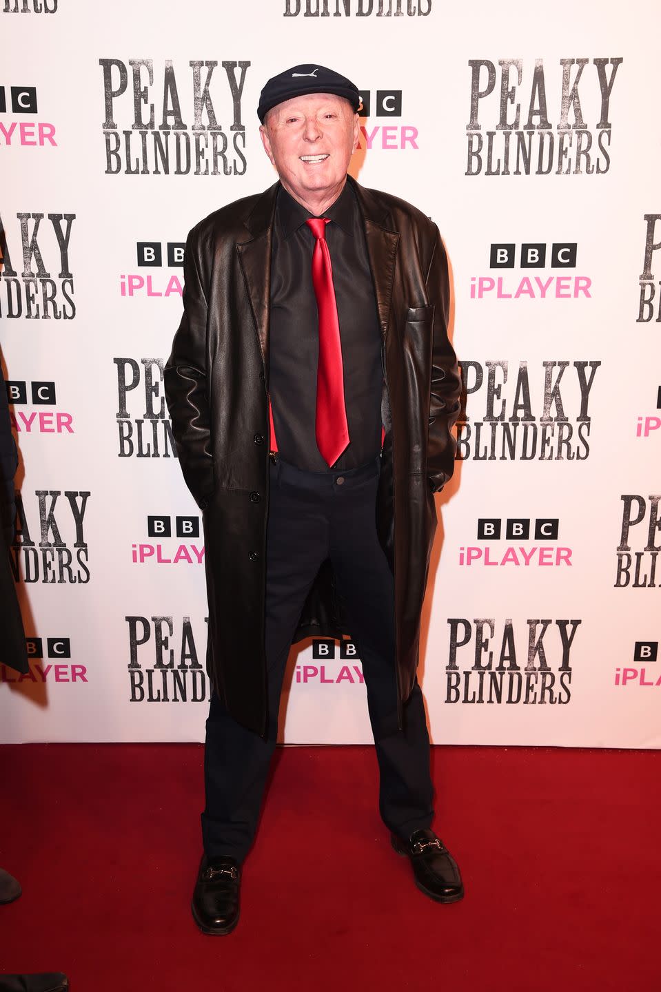 jasper carrott wearing a black jacket and red tie standing on a red carpet