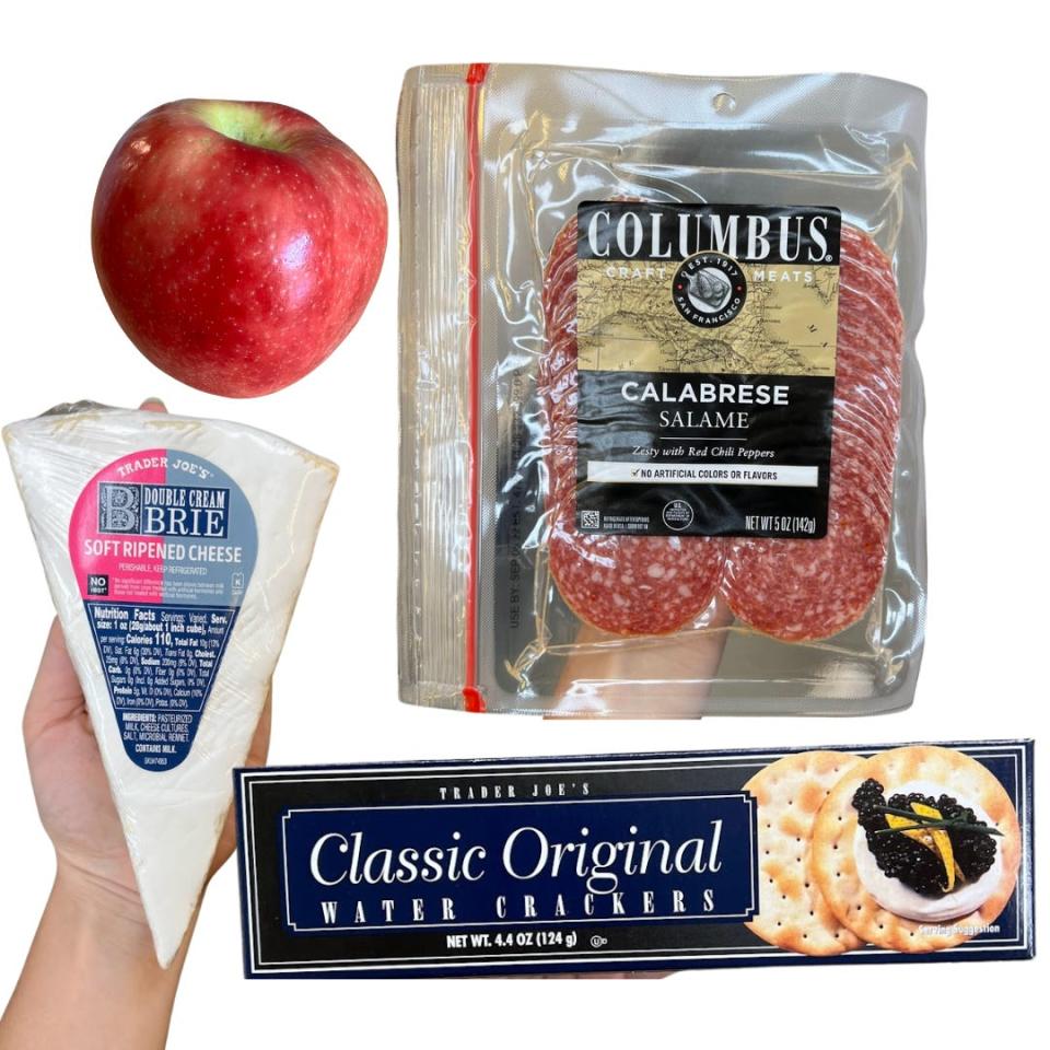 A red apple, a package of salami, a black box of crackers, and cheese held by the writer