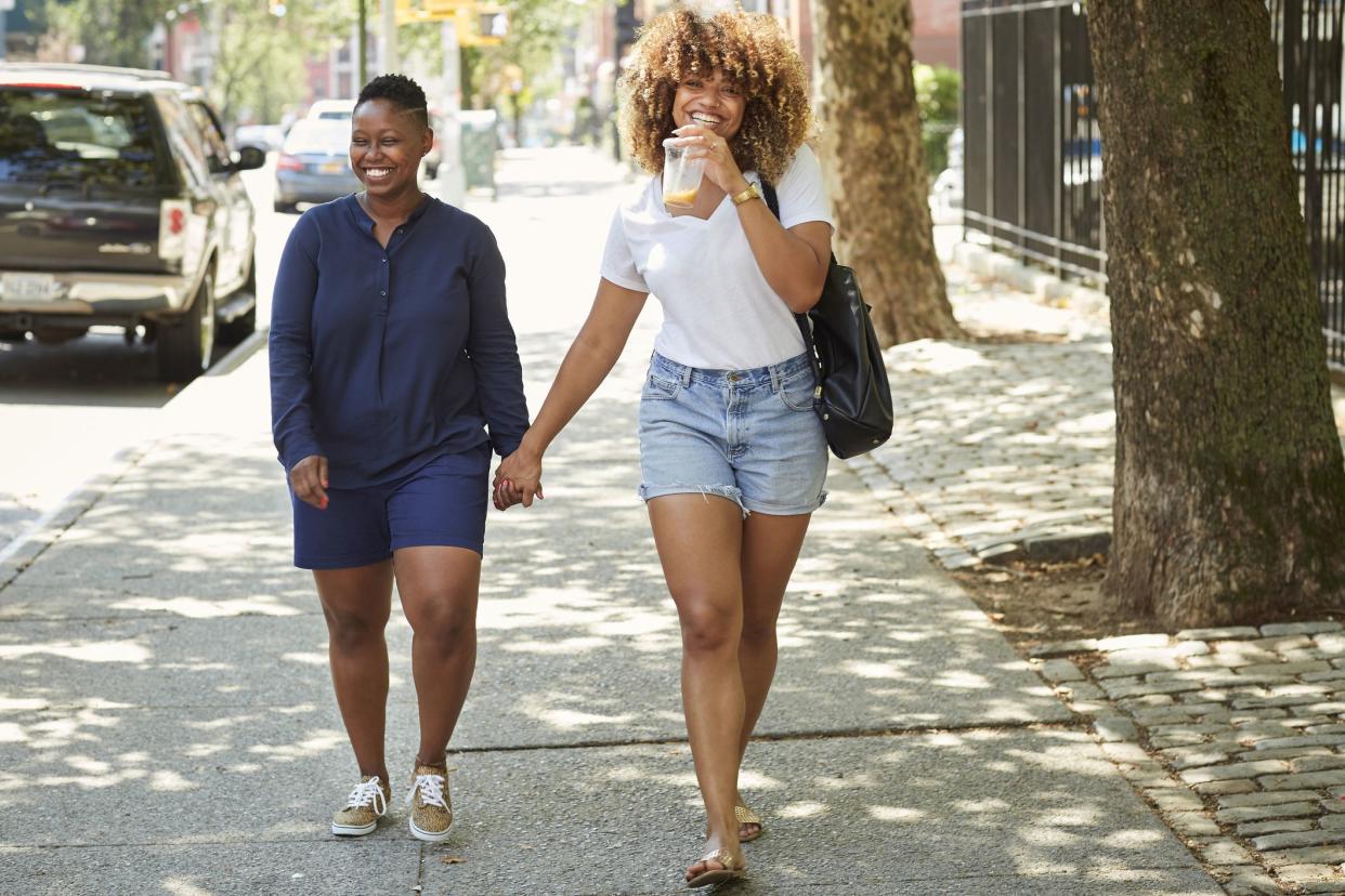 Young female couple walking on sidewalk, smiling, holding hands in a modern city, with buildings and cars in the background