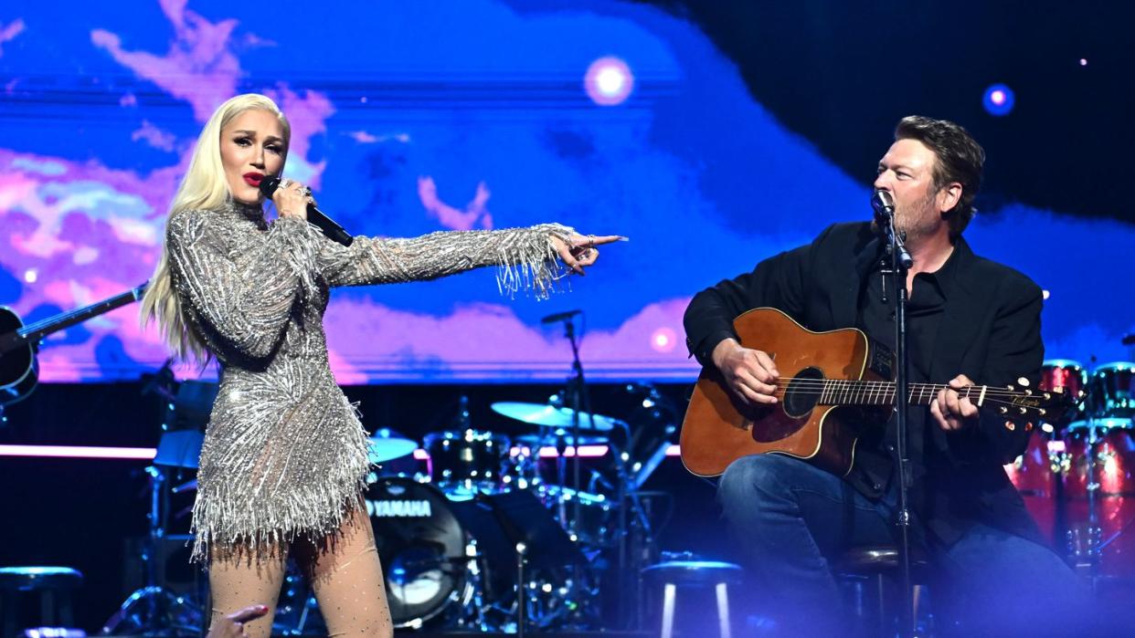 gwen stefani stands on a stage and sings into a wireless microphone she holds in one hand, blake shelton sits to the right as he plays guitar and sings into a microphone on a stand