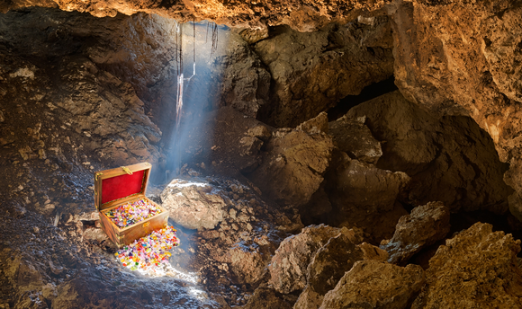 A treasure chest, trimmed in red velvet and spilling gold and jewelry over its edges, highlighted by a single ray of sunshine in an otherwise dark and barren cave.