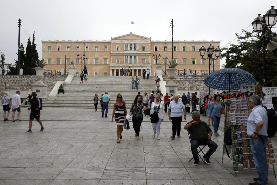 A lottery ticket vendor sits at his kiosk as pedestrians walk at Syntagma square in Athens, Monday, May 27, 2019. Greece's Prime Minister Alexis Tsipras called snap general elections following a resounding defeat of his left-wing Syriza party in European elections. The Greek Parliament is seen in the background. (AP Photo/Thanassis Stavrakis)