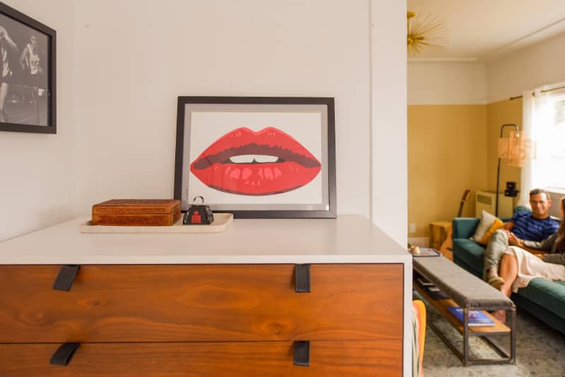 Artwork tops bedroom dresser as dweller is seen in living room.