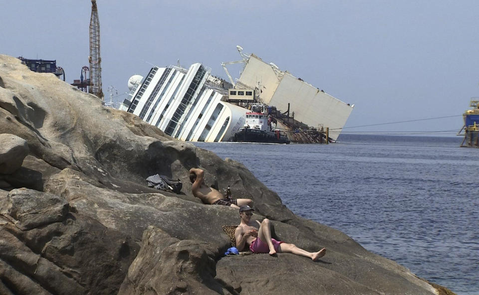 In this frame made from APTN video of Monday, July 8, 2013 made available Tuesday July 9, people sunbathe on the rocks as the wreckage of the Costa Concordia cruise ship is seen in the background, on the Tuscan island of Giglio. The trial of the captain of the shipwrecked Costa Concordia cruise liner began Tuesday in a theater converted into a courtroom in Grosseto, Italy, to accommodate all the survivors and relatives of the 32 victims who want to see justice carried out in the 2012 tragedy. The sole defendant, Schettino, made no comment to reporters as he arrived for his trial on charges of multiple manslaughter, abandoning ship and causing the shipwreck near the island of Giglio. His lawyer, Domenico Pepe, told reporters that, as expected, the judge was postponing the hearing though due to an eight-day nationwide lawyers' strike. Schettino has denied wrongdoing. (AP Photo via APTN)