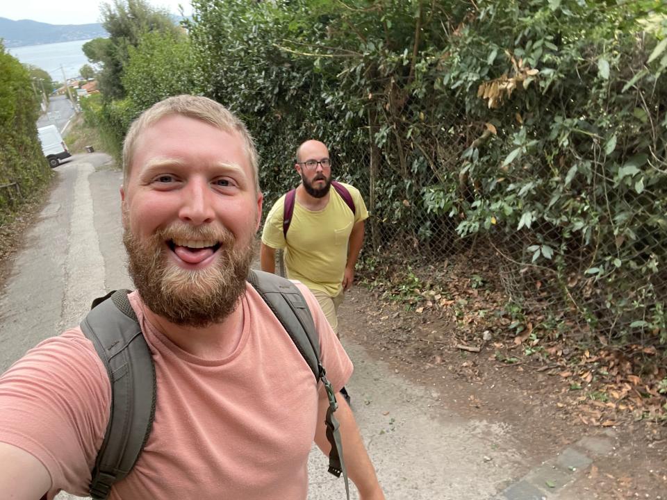 Selfie of the writer wearing a peach-colored shirt and sticking out his tongue while his husband wears a yellow shirt and walks behind him on a pathway in Italy