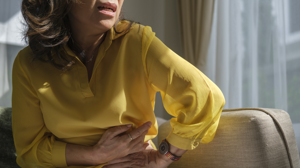 Older woman in long-sleeve yellow blouse touches her left side while grimacing in pain