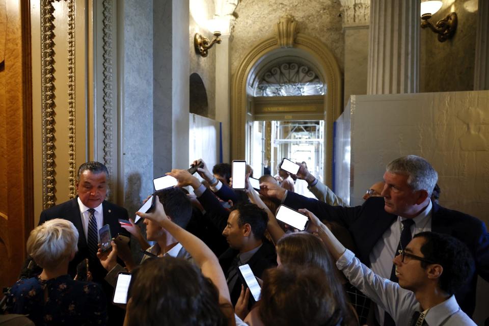 A man in a suit and tie talks in a high-ceilinged hallway to a large group of reporters recording him with their mobile phones.