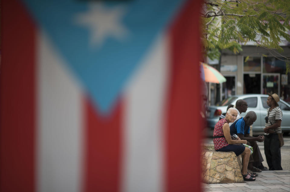 Residents sit outside the Plaza del Mercado in the Rio Piedras area of San Juan, Puerto Rico, Wednesday, April 17, 2019, where a Puerto Rican flag hangs. "You see fewer people around, fewer young people," said Wilfredo Montañez, 52, who was sitting on a bench in the plaza. (AP Photo/Carlos Giusti)