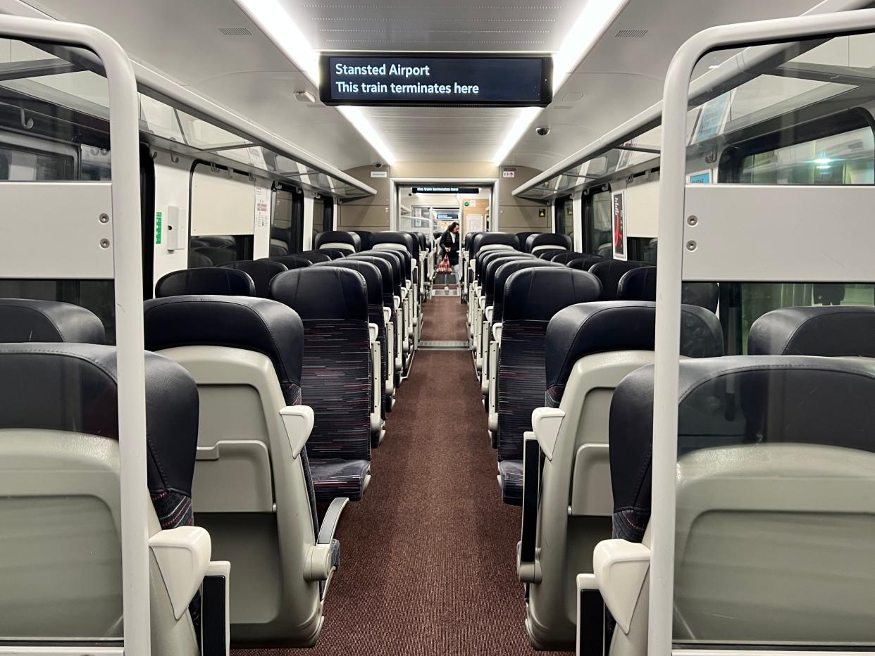 A view down the aisle of the Stansted Express train. Luggage racks are visible on the side, as well as several seats with black leather headrests and a moquette pattern
