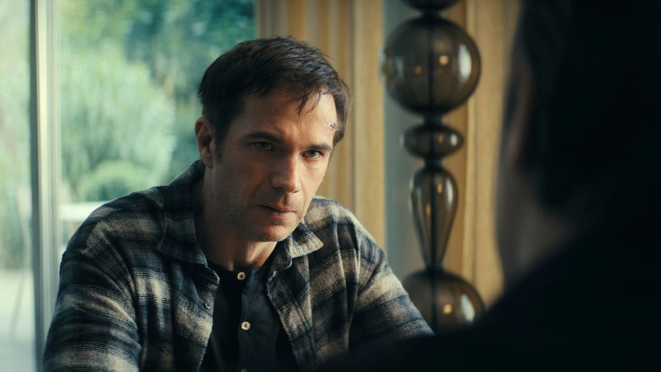 A man with short dark hair wearing a blue and gray plaid shirt is having a serious conversation with a person opposite him.