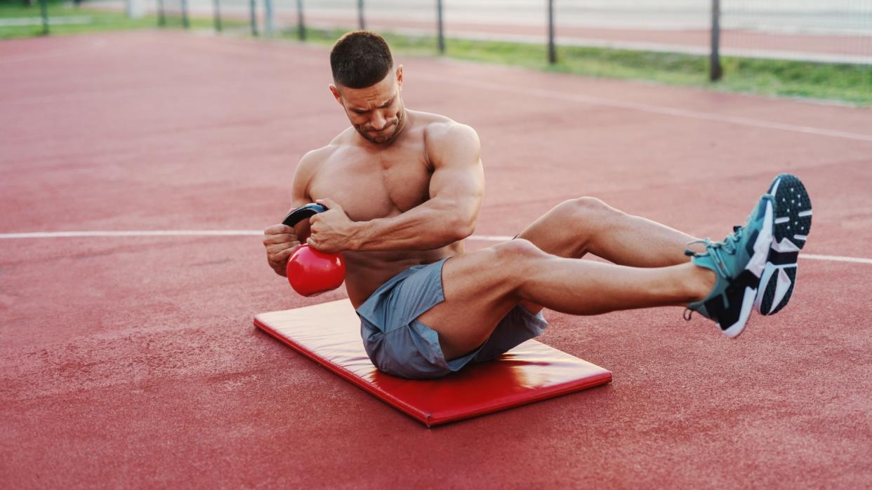  Man performing ab workout on mat outdoors showing a kettlebell Russian twist. 