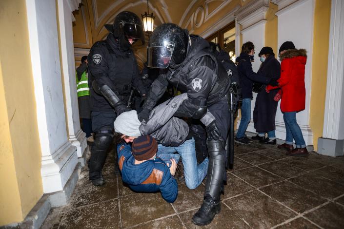 Police officers hold down two demonstrators in a building with classical Russian architecture.