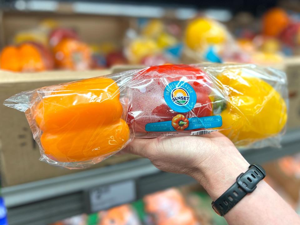 A hand holding a bag of orange, red, and yellow peppers.