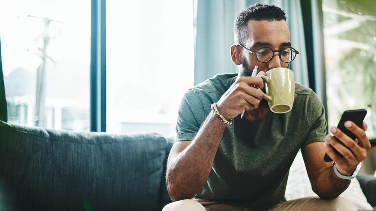 Shot of a young man using a smartphone and having coffee on the sofa at home.