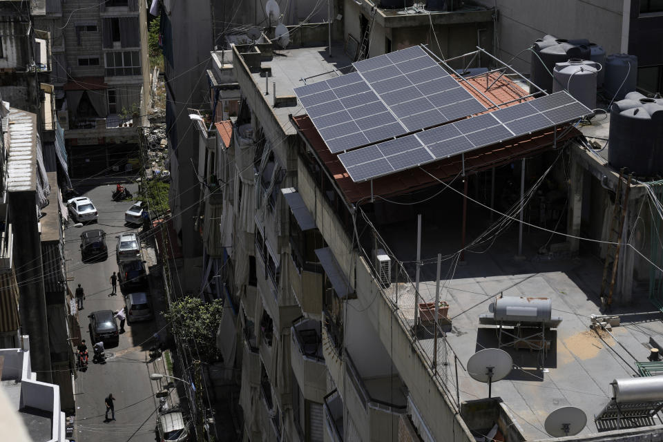 People walk in a street below solar panels installed over a building in Beirut, Lebanon, Thursday, May 30, 2024. Lebanese caretaker Minister of Economy and Trade Amin Salam said Lebanon's political class as well as fuel companies and private electricity providers in Lebanon blocked an offer by gas-rich Qatar to build three renewable energy power plants to ease the crisis-hit nation's decades-old electricity crisis. (AP Photo/Bilal Hussein)