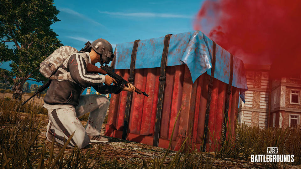 Still from PUBG, showing the player taking cover behind a crate with an assault rifle.  Village view with an old house in the background.