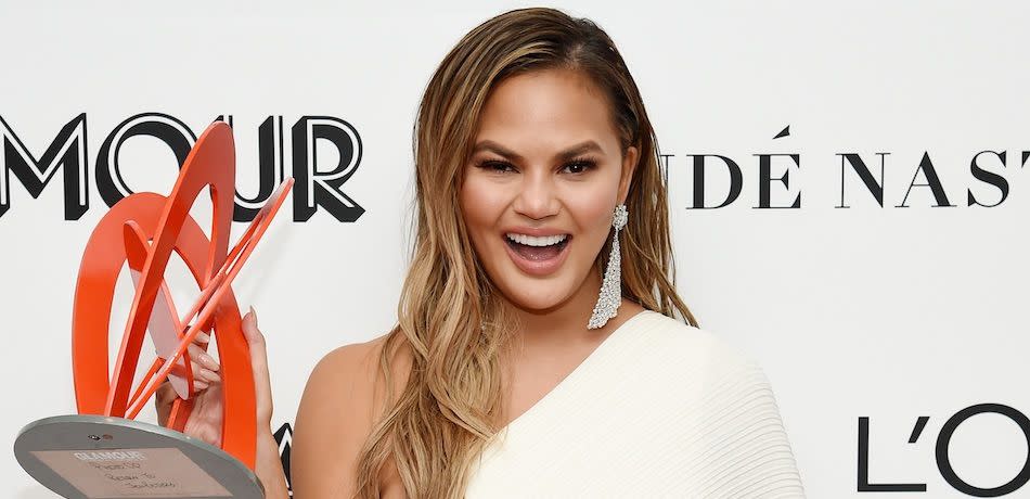 Chrissy Teigen grins while holding an award, wearing an off-the-shoulder white dress.