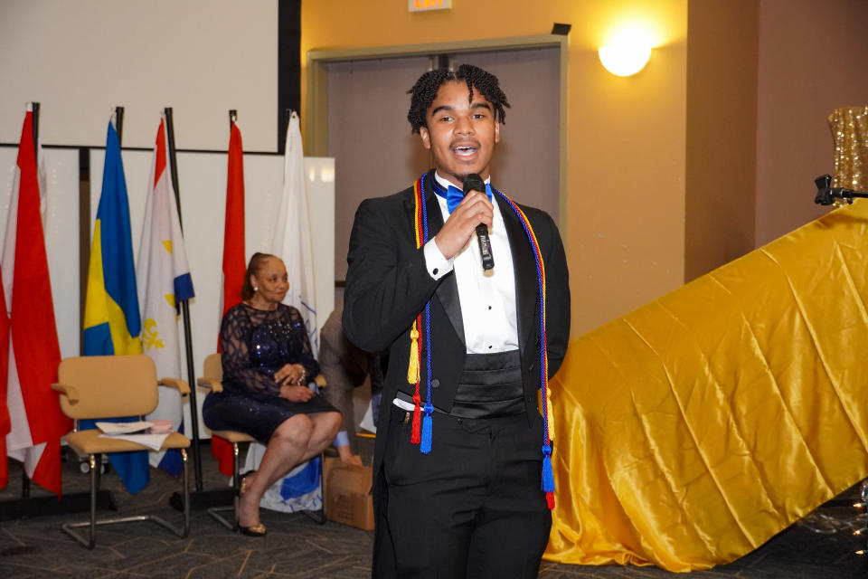 Dennis Barnes in tux and blue bow tie at the microphone in a school hall draped with flags and a yellow satin cloth.