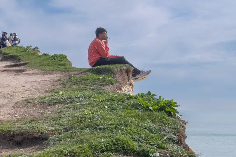 A man ignoring warning signs sitting on the edge of the cliff at The Seven Sisters National Park. A sightseer sits on a crumbly chalk 400 feet cliff casually puffing on a cigarette as he dangles his feet over the edge and dices with death.