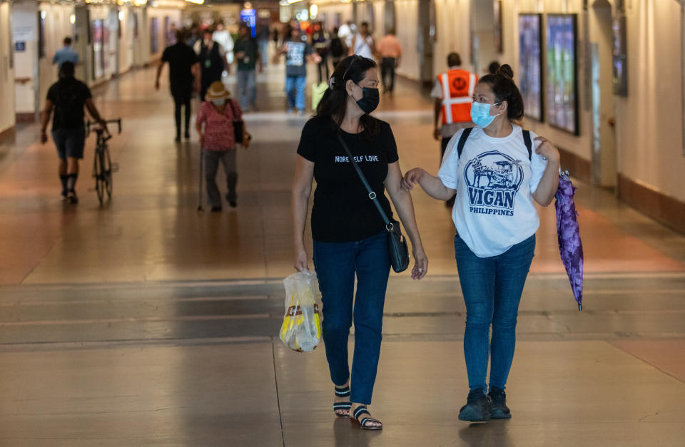 Two people wearing face masks walk together in a long, wide corridor with a few dozen people visible in the background.