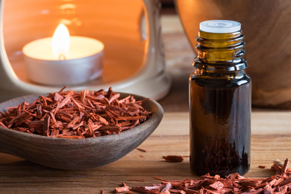 A bottle of sandalwood essential oil with sandalwood pieces and an aroma lamp in the background