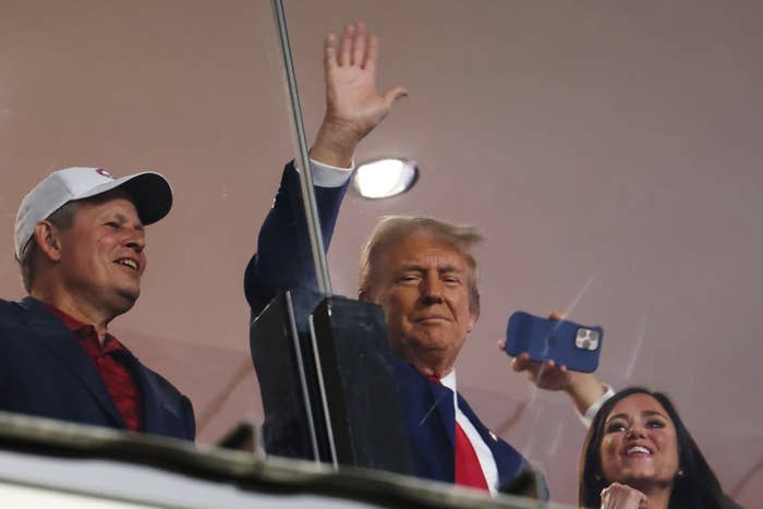 Donald Trump waves to the crowd in a suit and tie, accompanied by Sarah Huckabee Sanders, smiling and holding a phone, and another man in a cap