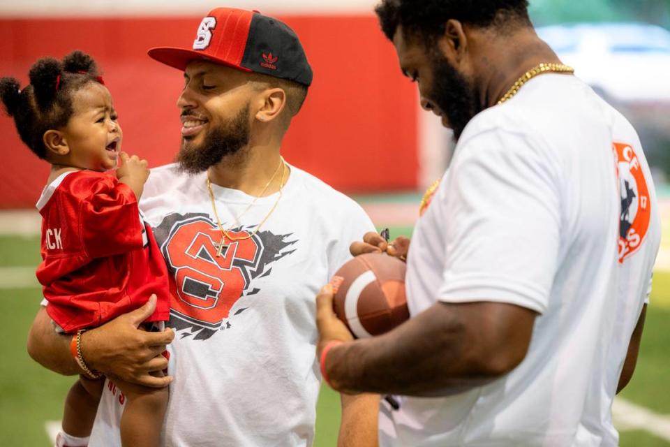 Jacoby Pulley holds his daughter Ivy Pulley, 1, as offensive lineman Jacarrius Peak autographs a football during N.C. State’s ‘Meet the Pack’ event with players, fans at the Close-King Indoor Practice Facility Saturday, Aug. 16, 2023.