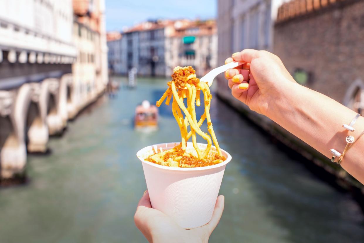 hand holding cup of pasta with fork pulling it out with venice canal in background