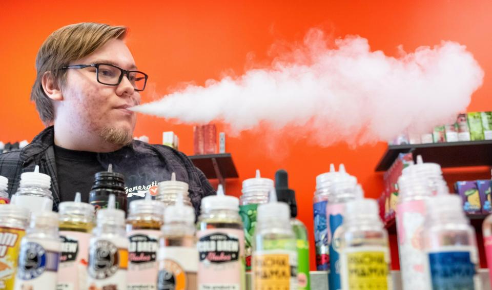 In this Nov., 2018 file photo, shift manager Shaun Miller exhales vapor while working behind the vape bar at Generation V in Omaha, Neb. An apparent surge in vaping among Nebraska teenagers is prompting a new push from lawmakers to raise the age limit on e-cigarettes from 18 to 21 and ban their use in bars, restaurants and workplaces. School officials say the crackdown would help them fight the growing use of e-cigarettes among young people.