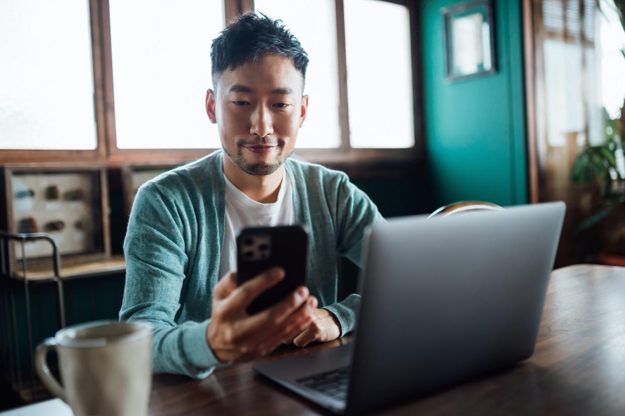 Confident young Asian man looking at smartphone while working on laptop computer in home office. Remote working, freelancer, small business concept