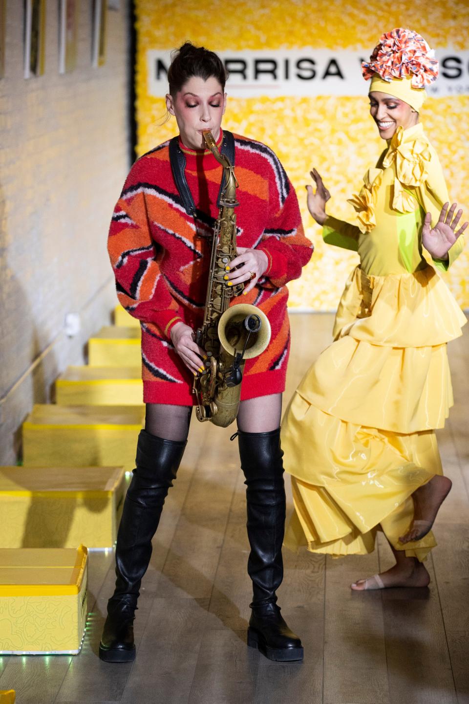 A woman plays the saxophone while a dancer dances down a runway.