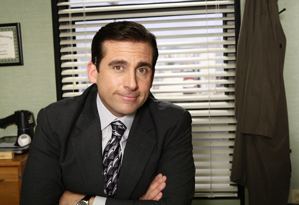 Steve Carell, dressed in a suit and tie, stands in an office with blinds behind him and a coat hanging to the right. He has a slight smile and arms crossed