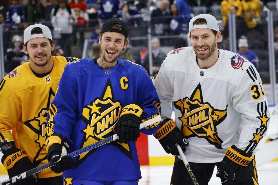 Three smiley men, in yellow, blue, and white hockey jerseys with NHL All-Star logos, pose together holding hockey sticks