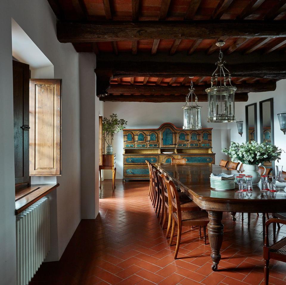 long view of a dining room with six or seven chairs at a table and two glass lantern pendant lights hanging from dark wooden beams over it and a zigzag terracotta floor