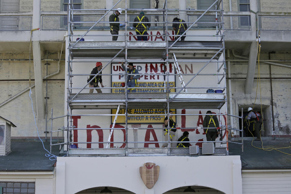 Eloy Martinez, on the center scaffolding, who took part in the original Native American occupation, waves while helping to repaint and restore messages painted above the main dock on Alcatraz Island Wednesday, Nov. 20, 2019, in San Francisco. About 150 people gathered at Alcatraz to mark the 50th anniversary of a takeover of the island by Native American activists. Original occupiers, friends, family and others assembled Wednesday morning for a program that included prayer, songs and speakers. They then headed to the dock to begin restoring messages painted by occupiers on a former barracks building. (AP Photo/Eric Risberg)