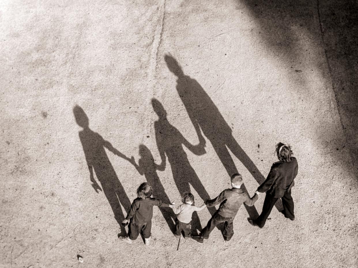 Three children and an adult hold hands and create four long shadows in front of them as they stand with their backs against the sun.