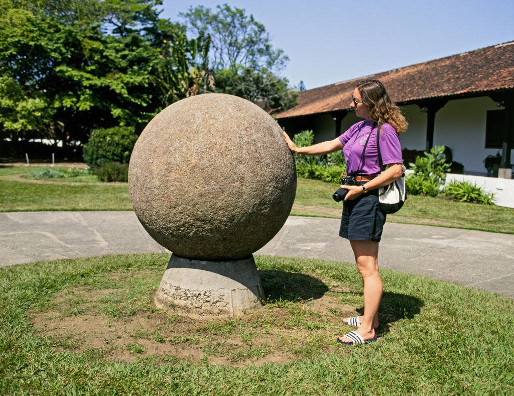 A woman in a purple shirt and black shirts touches a rock sphere that's almost as tall as her