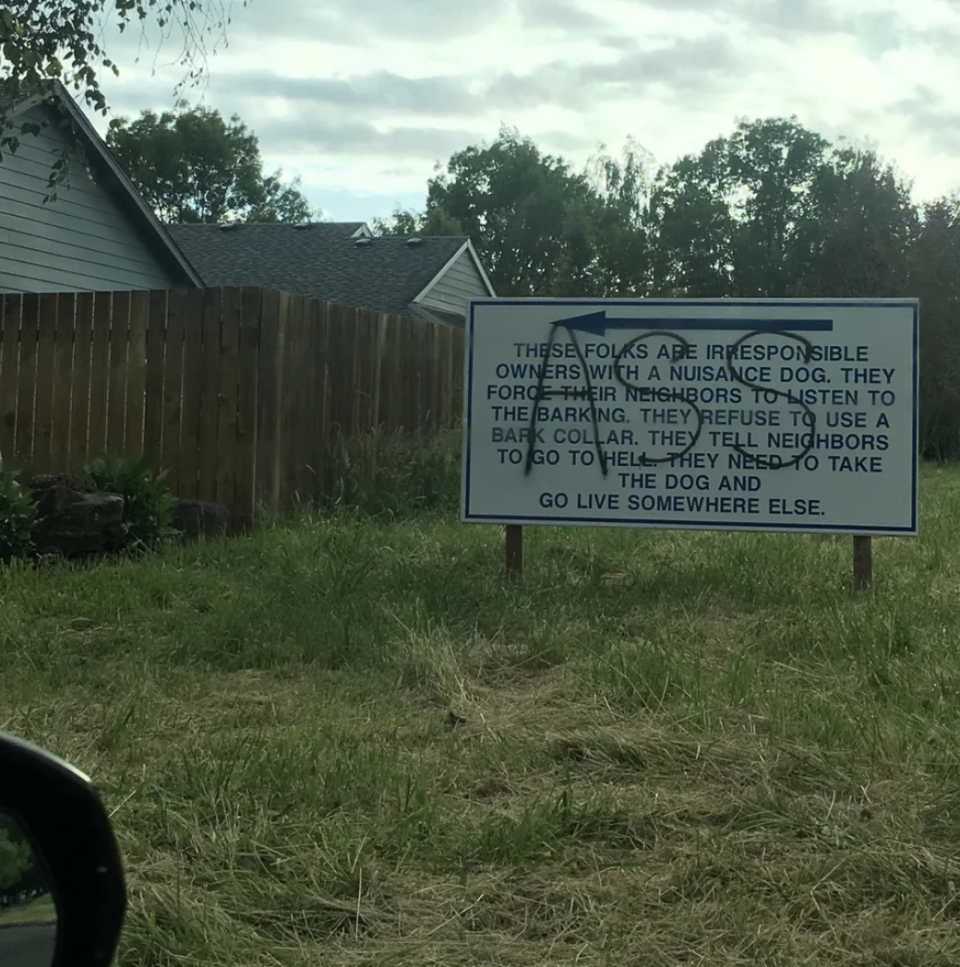 A large yard sign that says the people living next door are bad owners of a nuisance dog; the sign has been spray-painted with the word "ass" over the message