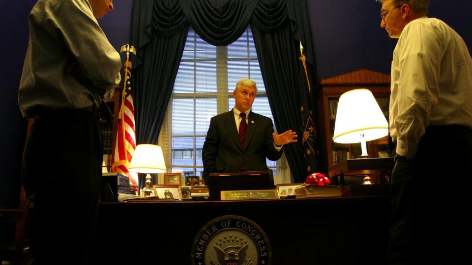 mike pence stands behind a desk with a congressional seal on the front, he looks at a man on the right of the desk as a man on the left side watches on, pence wears a suit with a white shirt and red tie, behind him is a window with ornate curtains and two flags