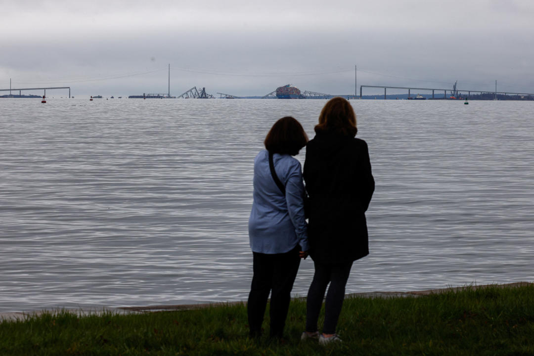 Two people standing on a shoreline, seen from behind, face the collapsed Francis Scott Key Bridge.