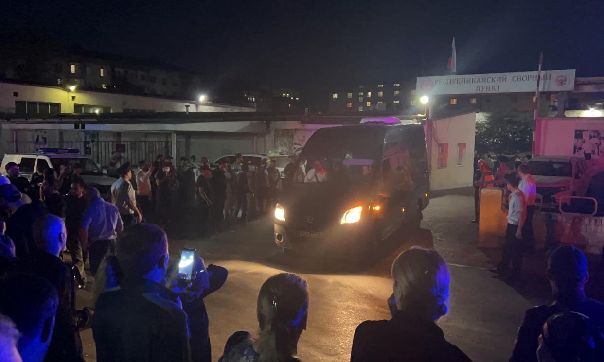 A group of people on a street at night watch a truck drive away.