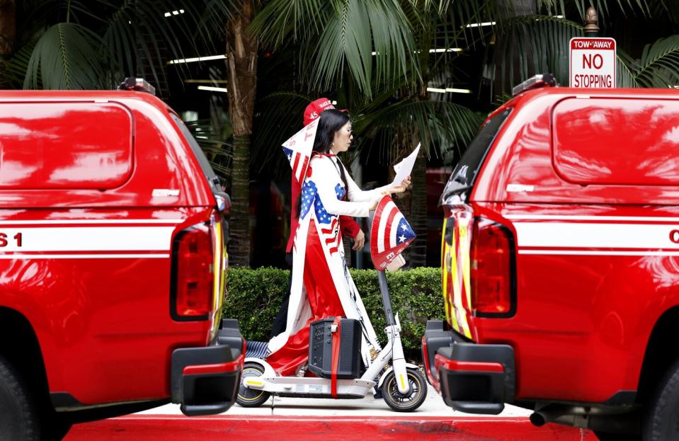 A woman in red, white and blue seen in the gap between two red vehicles as she rolls by with a speaker strapped to a scooter