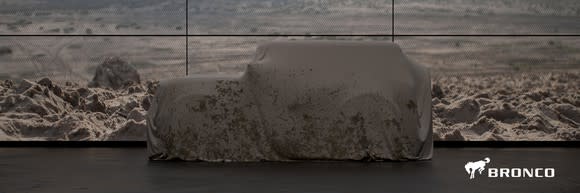 An angular SUV is shown under a cover in front of a rocky background