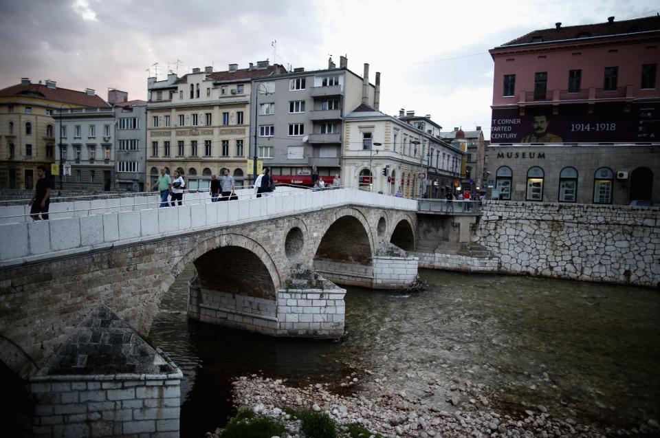 People cross the Latin Bridge and street corner in front of the historical landmark, where Archduke Franz Ferdinand and his wife Sophie were assassinated, in Sarajevo June 24, 2014. Two concerts in two Bosnian cities, Sarajevo and Visegrad respectively, will mark the 100th anniversary on June 28, 2014 of the Sarajevo assassination of Ferdinand that lit the fuse for World War One, in a divided country where the past still haunts the present.Picture taken June 24, 2014. To match WWI-ANNIVERSARY/BOSNIA. REUTERS/Dado Ruvic (BOSNIA AND HERZEGOVINA - Tags: CONFLICT ANNIVERSARY SOCIETY POLITICS)