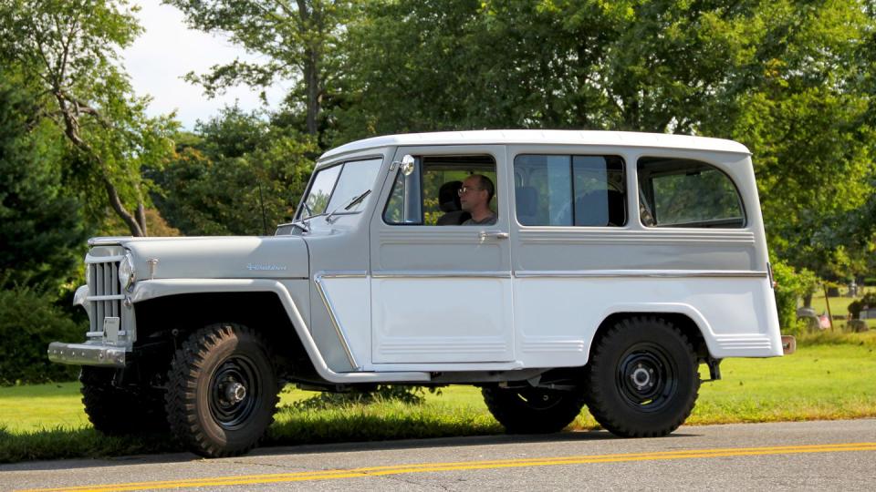 a white and grey 1962 willys jeep station wagon sits on the side of a road with trees in the background