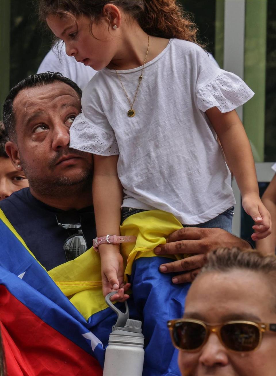 Davie resident Victor Macedo, 39, left, looks towards his daughter Sonia, 5, perched on his shoulder after the signing of the Venezuelan National Anthem during the demonstration for the right to vote in the Venezuelan election. A group of about sixty people gathered at the former Venezuelan consulate in protest of their voting denial in the Venezuelan election on Sunday, July 28, 2024, in Miami, Florida.